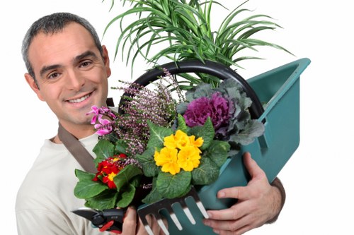 A professional gardener holding a green plastic watering can filled with a variety of freshly potted flowering plants and green foliage, with a young, smiling man dressed in a light-colored shirt. The garden tools and plants are set against a neutral background, highlighting the vibrant yellow, purple, and red blossoms, as well as the lush green leaves. The scene suggests a well-maintained outdoor space, possibly part of a landscaped front or back garden in Swiss Cottage, with the gardener ready to carry out planting or watering tasks. The image reflects attention to gardening and outdoor care, consistent with services offered by Swiss Cottage Gardening Services, emphasizing quality lawn and plant care in an urban outdoor environment under natural lighting conditions.