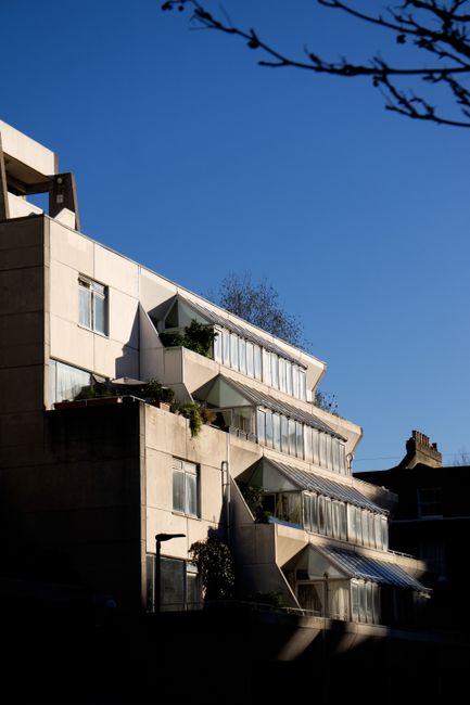 A multi-storey residential building with a modern architectural design featuring multiple balconies with glass railings, some adorned with potted plants and small shrubs. The building has an angular s