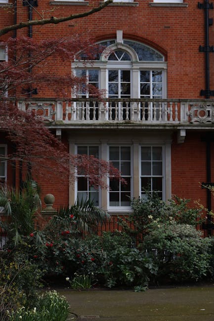 A multi-storey residential building with a modern architectural design featuring multiple balconies with glass railings, some adorned with potted plants and small shrubs. The building has an angular s